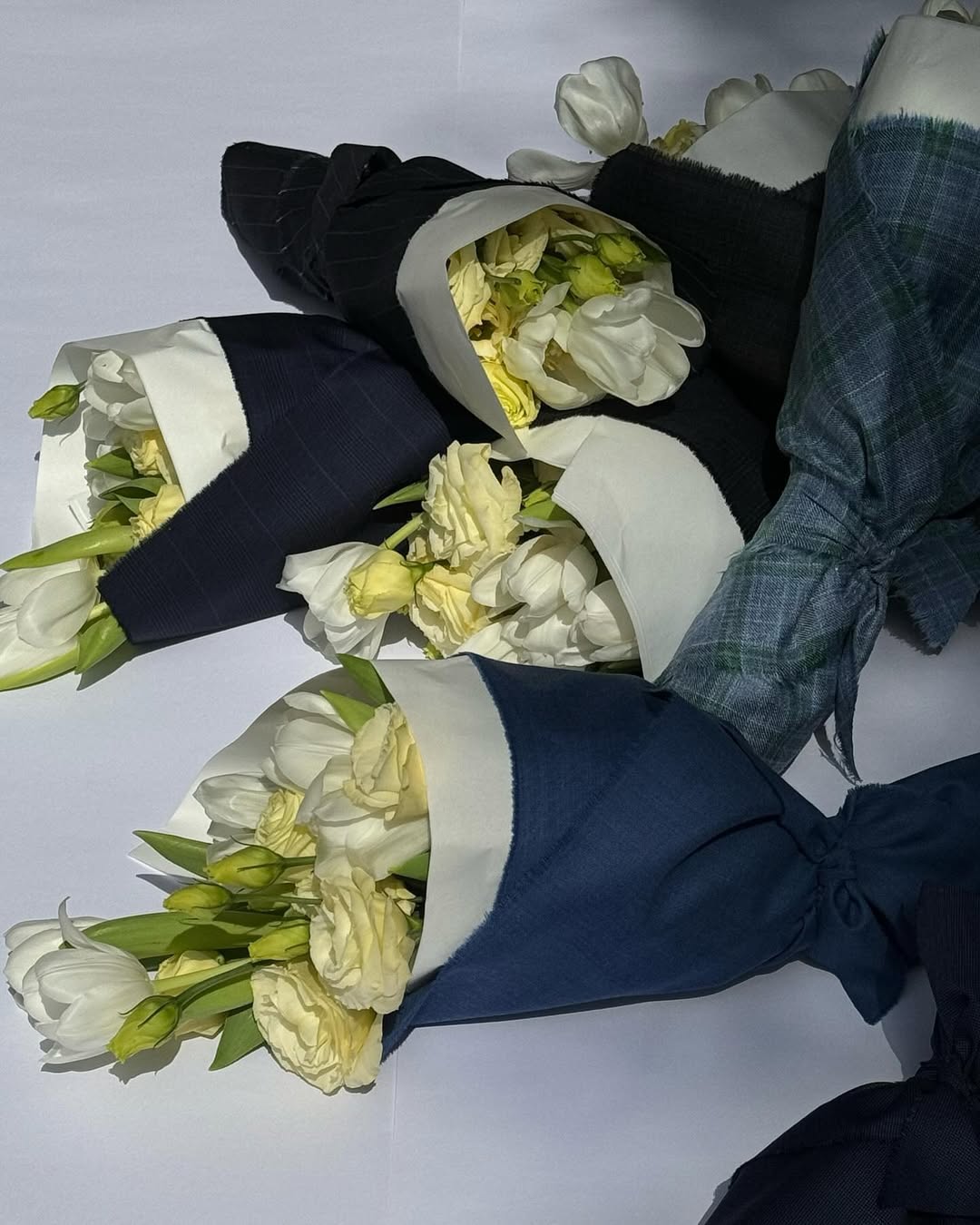 Buquês de tulipas brancas e flores em tom creme, envolvidos em tecidos azul-marinho, preto e azul acinzentado.