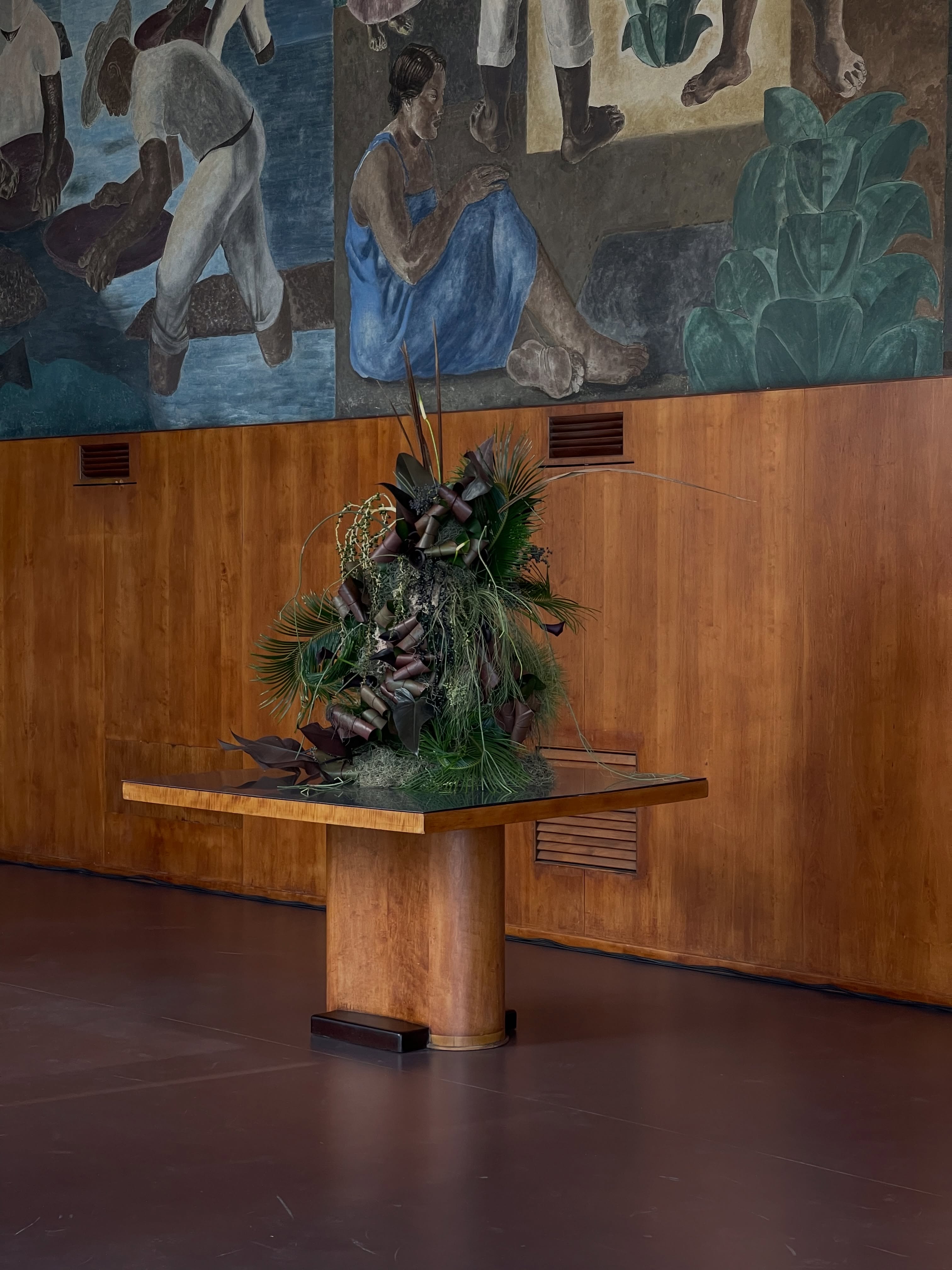 Escultura botânica sobre mesa de madeira em um espaço com painel de arte.
